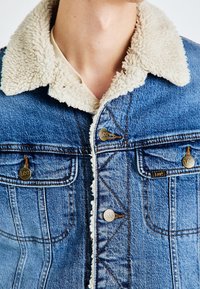 Jeansjacka med en beige sherpa-krage, mässingsknappar och bröstfickor. Blå denimtyg med subtila sömnadsmönster.