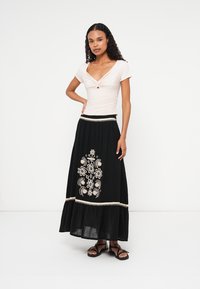 Svetlo roza prilegajoča majica z zasukanim vozlom, v kombinaciji z črno maksi krilo s belim cvetličnim vezenjem in plastenim robom.