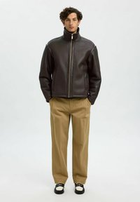 Brun läderjacka med hög krage och dragkedja framtill, kombinerad med beige byxor och svart-vita skor. Slät textur, minimalistisk design.