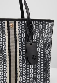 Sac cabas à motif noir et blanc avec des rayures verticales, anses en cuir noir et une étiquette noire avec un symbole de logo.