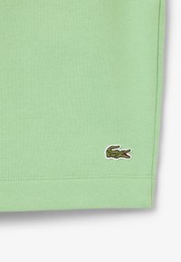 Tissu en coton vert clair avec une texture lisse. Présente un petit logo de crocodile brodé en vert et blanc près de l'ourlet.
