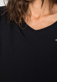 Černé tričko s krátkým rukávem a výstřihem do V vyrobené z měkkého bavlněného materiálu, s malým bílým logem na levé straně poblíž výstřihu.