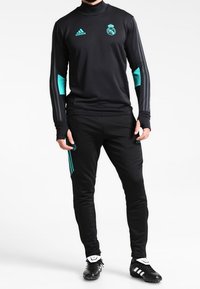 Mann in schwarzem Real Madrid Trainingsanzug mit türkisfarbenen Adidas-Logos und -Streifen, der vor einem weißen Hintergrund steht.