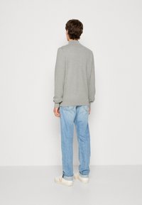 En man står med ryggen mot kameran, iförd en grå tröja, ljusblå jeans och vita sneakers, framför en helt vit bakgrund.