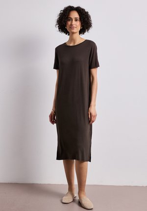 Femme aux cheveux bouclés portant une robe midi marron foncé à manches courtes et des chaussures beige sans lacets, debout contre un mur clair uni.