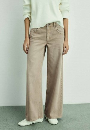 Person iført beige jeans med vide ben, hvide sneakers og en cremefarvet ribstrikket sweater stående mod en ensfarvet lys baggrund.