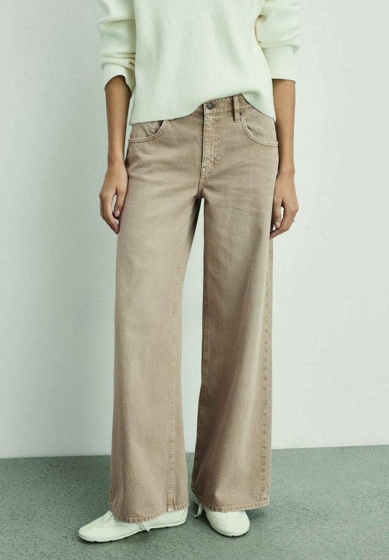 Personne portant un jean beige à jambes larges, des baskets blanches et un pull côtelé crème, debout devant un fond clair uni.