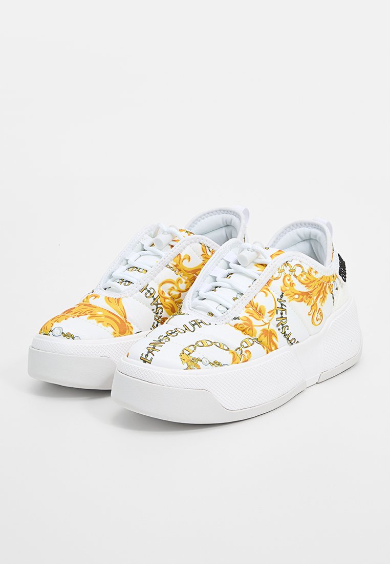 Weiße Sneaker mit glänzender Oberfläche, verziert mit gelben ornamentalen Mustern und bedrucktem Text. Dicke Gummisohle mit strukturierten Akzenten. Klassisches Design.