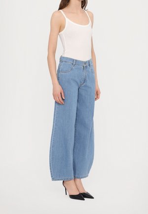 Vrouw draagt lichtblauwe, hoog getailleerde jeans met wijde pijpen, wit mouwloos topje en zwarte spitse hoge hakken, staand tegen een witte achtergrond.