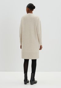 Cardigan en tricot beige avec des manches longues, des poignets et un ourlet côtelés, d'une longueur jusqu'aux genoux, présentant une texture lisse et des coutures minimales.