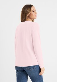 Helles rosafarbenes Langarmshirt mit Rundhalsausschnitt und schlichten Design. Weiche Textur, minimales Branding auf der Rückseite, kombiniert mit blauen Jeans.