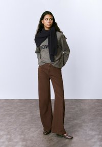 Mujer de pie, vestida con un suéter gris con texto, una bufanda oscura caída sobre los hombros, pantalones anchos marrones y zapatos oscuros con tachuelas, sobre un fondo liso.