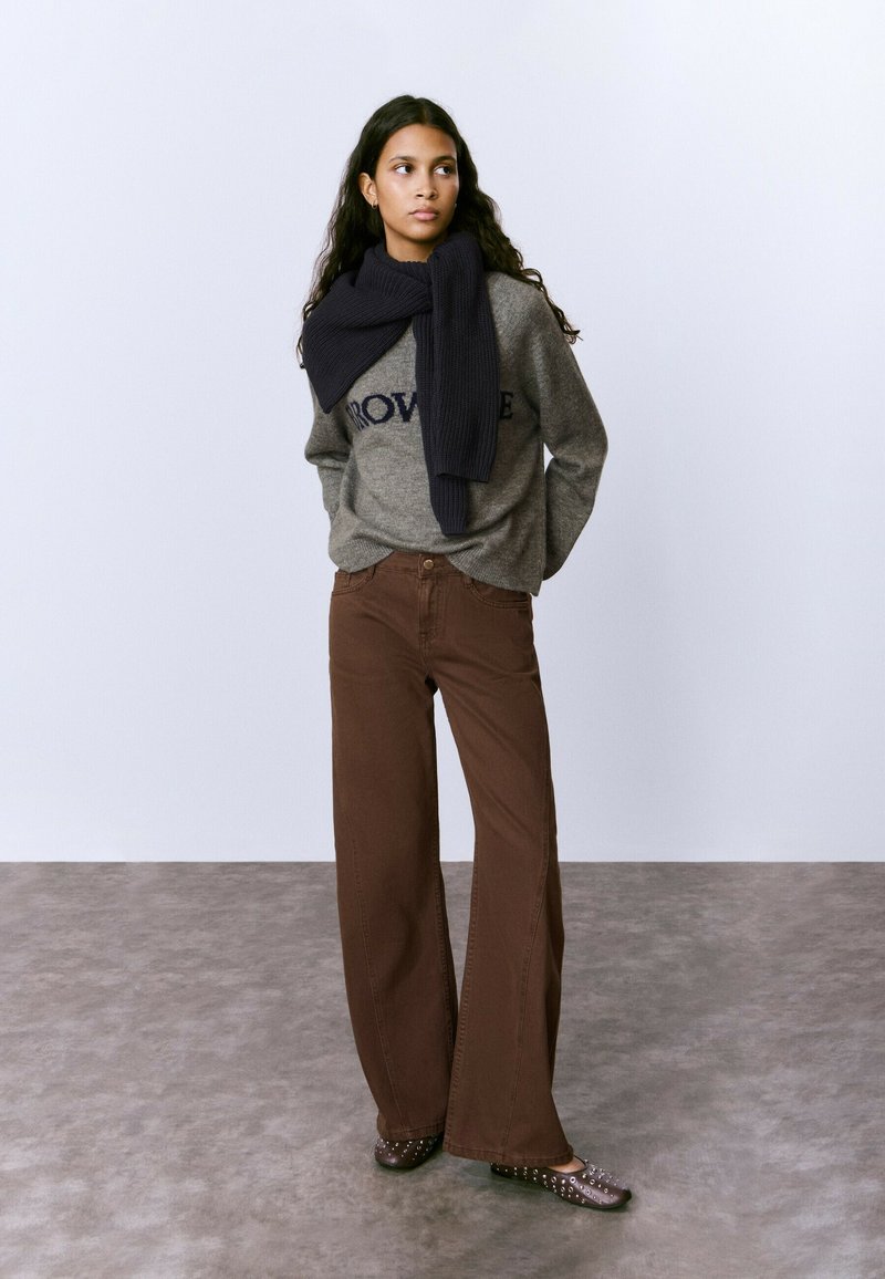 Mujer de pie, vestida con un suéter gris con texto, una bufanda oscura caída sobre los hombros, pantalones anchos marrones y zapatos oscuros con tachuelas, sobre un fondo liso.