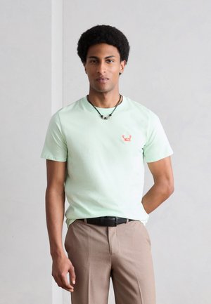 Mężczyzna z krótkimi kręconymi włosami, ubrany w jasniefioletową koszulkę z małym czerwonym logo kraba, beżowe spodnie i czarny naszyjnik, stojący przy szarej ścianie.