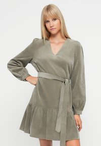 Olivengrønn kjole i myk stoff, med V-hals, puffede ermer, en tettsittende midje med belte og en utsvakt skjørt.