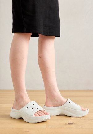 Λευκά καουτσούκ ξυλοπόδαρα με αεραγωγούς, στρογγυλή σχεδίαση, χοντρή σόλα και υφή στην κάτω επιφάνεια, φορεμένα με μια μαύρη φούστα μέχρι το γόνατο.