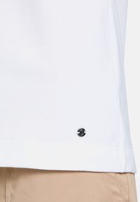 T-shirt en coton blanc avec un col ras du cou et un accent décoratif en bouton noir près de l'ourlet. Texture lisse et coupe décontractée.