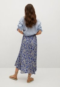 Gonna maxi a fantasia paisley blu con un design fluido, abbinata a una camicia button-down azzurro chiaro e scarpe beige.