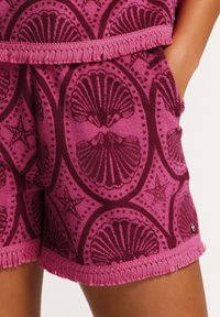 Fleece šortky v živé růžové barvě s motivem mušlí, s texturovaným povrchem a třásněmi na lemu. Ozdobené detailem loga.