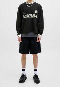 Felpa nera con accenti bianchi, caratterizzata dalla grafica "VENTURA". Abbinata a pantaloncini cargo neri, calzini bianchi con strisce nere e scarpe nere.