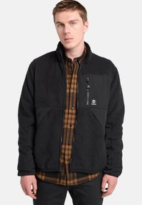 Giacca in pile nera con zip frontale, pannelli in nylon neri a contrasto e patch con logo. Indossata sopra una camicia a quadri marrone.