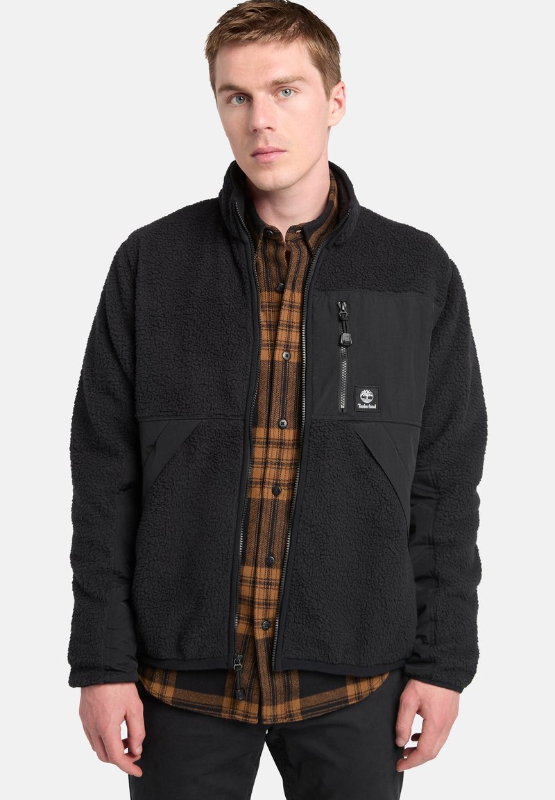 Giacca in pile nera con zip frontale, pannelli in nylon neri a contrasto e patch con logo. Indossata sopra una camicia a quadri marrone.