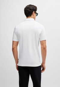 White short-sleeve polo shirt made of cotton, featuring a classic collar and side slits. Worn with black pants and sunglasses.