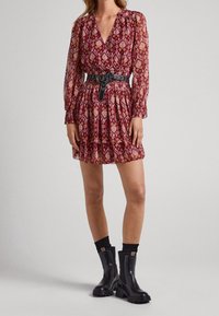 Rotes gemustertes Kleid mit V-Ausschnitt, elastischem Bund und Ballonärmeln, kombiniert mit schwarzen Knöchelboots. Das Kleid weist aufwendige Diamantmuster auf.