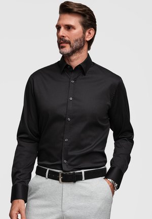 Homme portant une chemise noire cintrée boutonnée, rentrée dans un pantalon gris clair avec une ceinture noire, debout avec une main dans la poche.