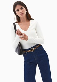 Joven mujer que lleva una blusa de manga larga en color blanco sucio, jeans de tiro alto azul oscuro con un cinturón de estampado de leopardo, sosteniendo un bolso negro de hombro, contra un fondo blanco.