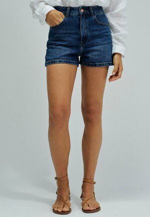 Mujer con shorts vaqueros azul oscuro, camisa blanca de manga larga y sandalias planas marrones con tiras, de pie con una mano en el bolsillo.