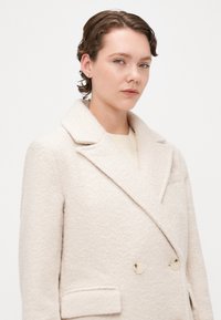 Lys beige dobbeltradet frakk, laget av teksturert stoff, med spiss revers, to frontlommer og kremfargede knapper.