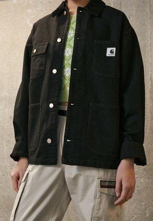 Sort denimjakke med krave, to forlommer, trykknapper og et logo-patch; båret over en grøn blomstret striktrøje og beige bukser.