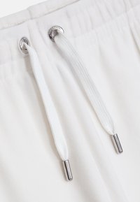 Λευκές αθλητικές σορτς με μαλακή υφή, ελαστική μέση, και ασημένια οπές με λευκές κορδόνες που έχουν μεταλλικά άκρα.