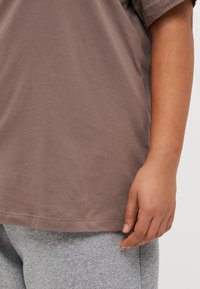 Rudas medvilninis marškinėlis su apvaliu kaklu, derinamas su šviesiai pilkomis kelnėmis. Matoma ranka, atsirėmusi į marškinėlių šoną.