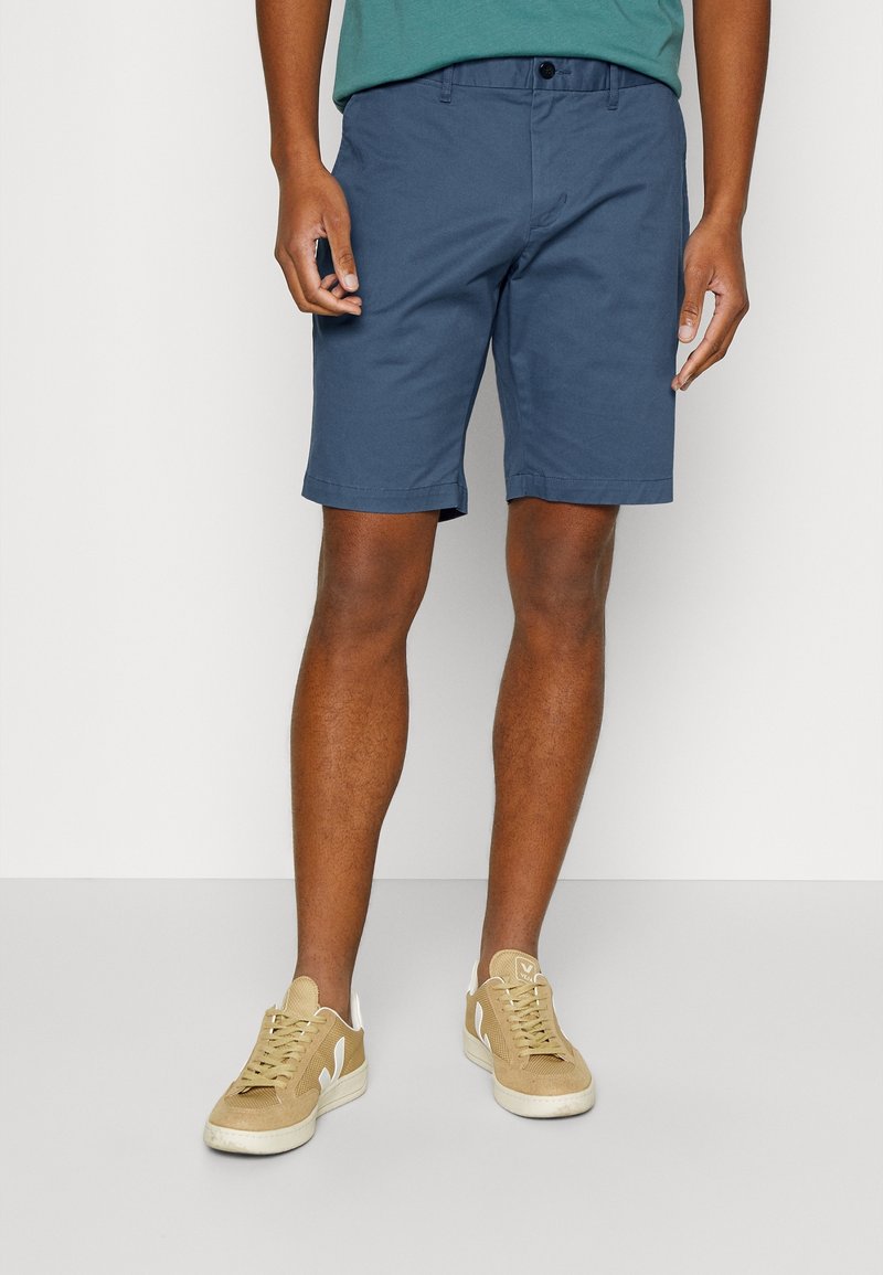 Homme portant un short bleu au genou, une chemise bleu sarcelle et des baskets beiges, debout devant un fond clair uni.