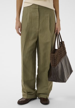 Osoba nosiaca voľné olivovo zelené nohavice a hnedé mokasíny, držiaca veľkú hnedú prepletenú kabelku z kože a látky.