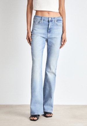 Vrouw draagt lichtblauwe hooggetailleerde flare jeans met witte cropped top en zwarte open-toe sandalen met hakken, staand op een lichte vloer.