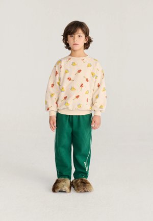 Enfant aux cheveux bruns debout portant un sweat-shirt beige avec des imprimés de champignons jaunes et rouges, un pantalon vert et de grandes chaussons en fourrure.