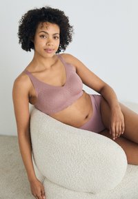 Mauve bralette dengan pola tekstur dan tepi bergelombang, dipadukan dengan celana dalam tinggi yang cocok di kursi krem bertekstur.
