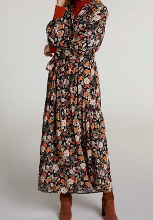 Femme portant une longue robe noire à motifs floraux orange et blancs, avec des bottines en suède marron.