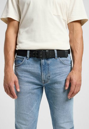 Cinturón negro de tela con hebilla de metal, llevado sobre unos jeans de mezclilla azul claro. El cinturón cuenta con múltiples agujeros de ajuste y un acabado texturizado.