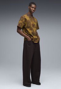 Korte mouwen polo in olijfgroen met een paisleypatroon; met een bruine kraag en loszittende donkere broeken met wijde pijpen.