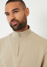 Sweatshirt zippé beige clair avec un col montant et une fermeture éclair en argent. Texture douce et lisse avec des détails de couture minimalistes.