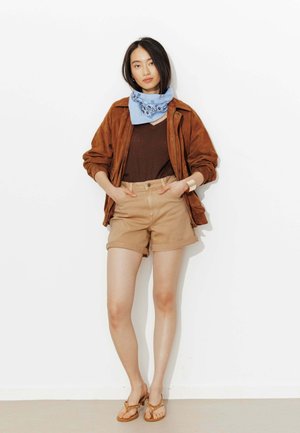 Femme debout contre un mur blanc, portant une veste marron, un haut marron, un short beige, un foulard bleu à motifs autour du cou et des sandales beige, les mains dans les poches.