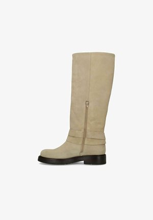 Beige suède laarzen tot aan de knie met ritssluiting aan de zijkant, decoratieve riempjes boven een platte zwarte zool met lage hak.