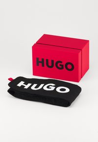Bandana negra con el texto blanco "HUGO", hecha de tela suave, colocada al lado de una caja rectangular roja con la misma marca.