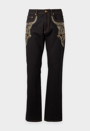 Modello maschile che indossa una maglietta rosa senza maniche con rifinitura marrone scuro, pantaloni neri con ricamo paisley dorato e scarpe nere con punte dorate.