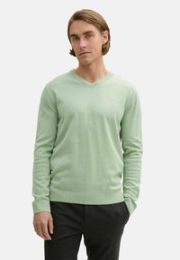 Pulover verde deschis cu decolteu în V, cu mâneci lungi, textură moale și manșete cu nervuri. Purtați peste pantaloni închiși, având un detaliu subtil cu logo.