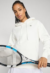 Hvit hettegenser med liten grønn Lacoste-logo, båret av en person som holder en blå og svart tennisracket, satt mot en nøytral bakgrunn.